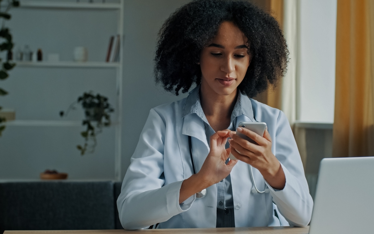 A doctor in a white coat using a smartphone at a desk with a laptop to verify AI output.